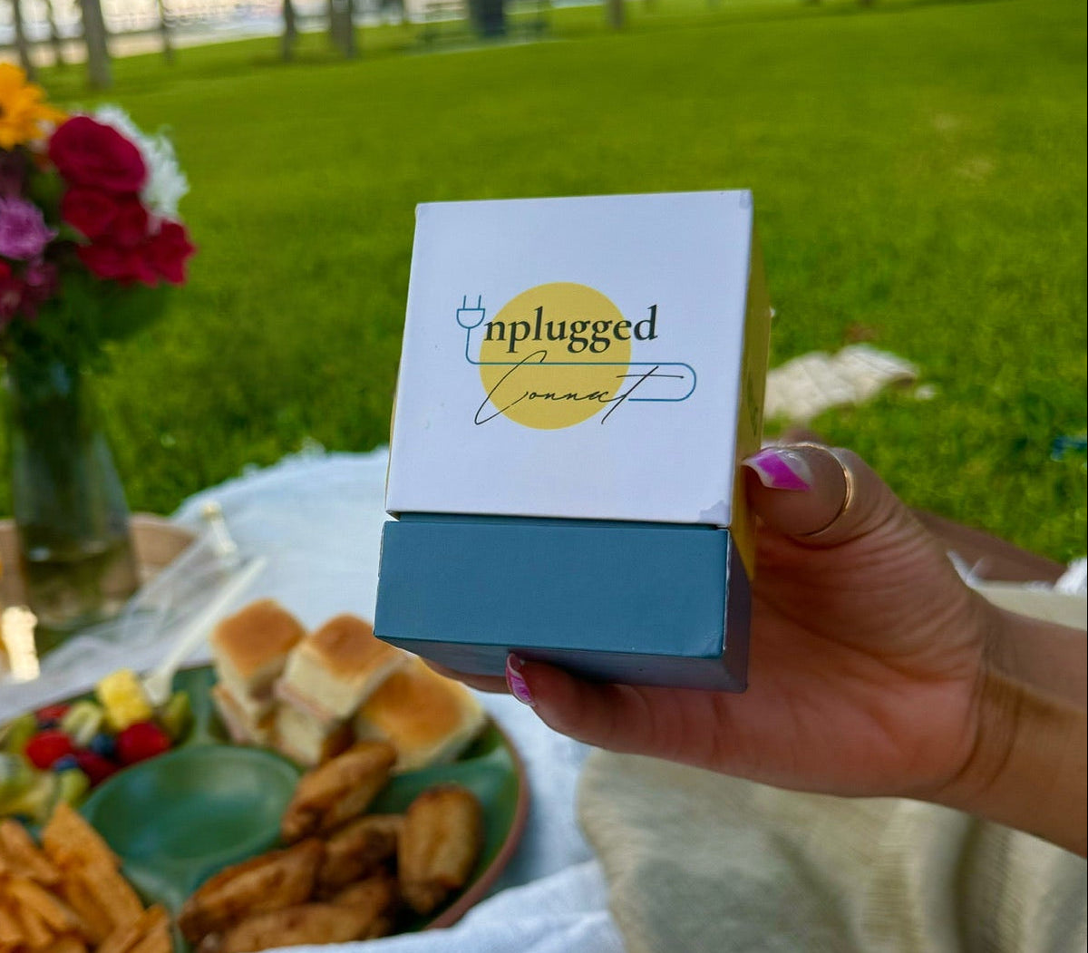 Person holding a 'plugged in' product at a picnic with food and flowers.