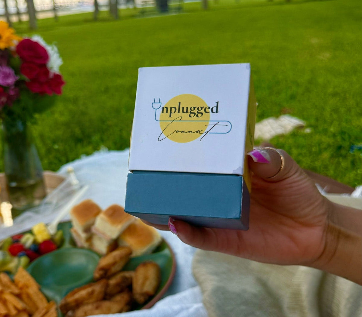 Person holding a 'plugged in' product at a picnic with food and flowers.