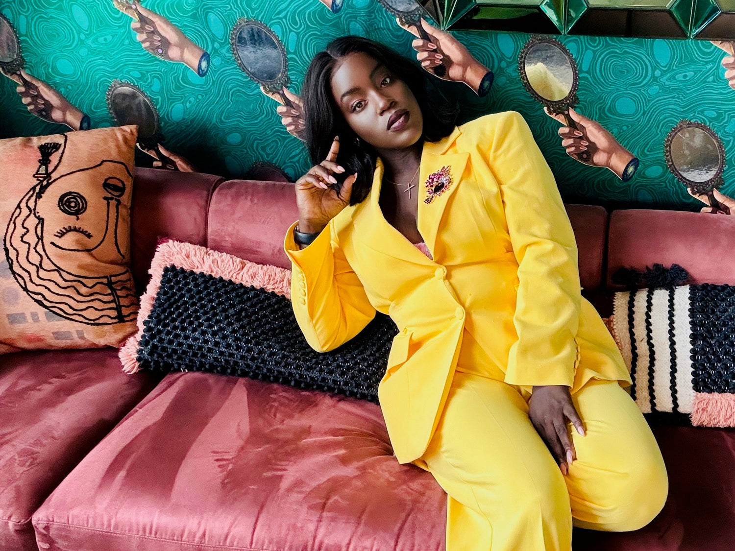 Woman in a yellow outfit sitting on a pink couch with a colorful wall and mirror reflections.

